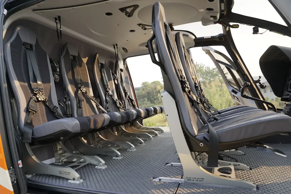 Interior of a modern helicopter showing two rows of seats with four-point harness seatbelts. The helicopter has a spacious cabin with large windows.