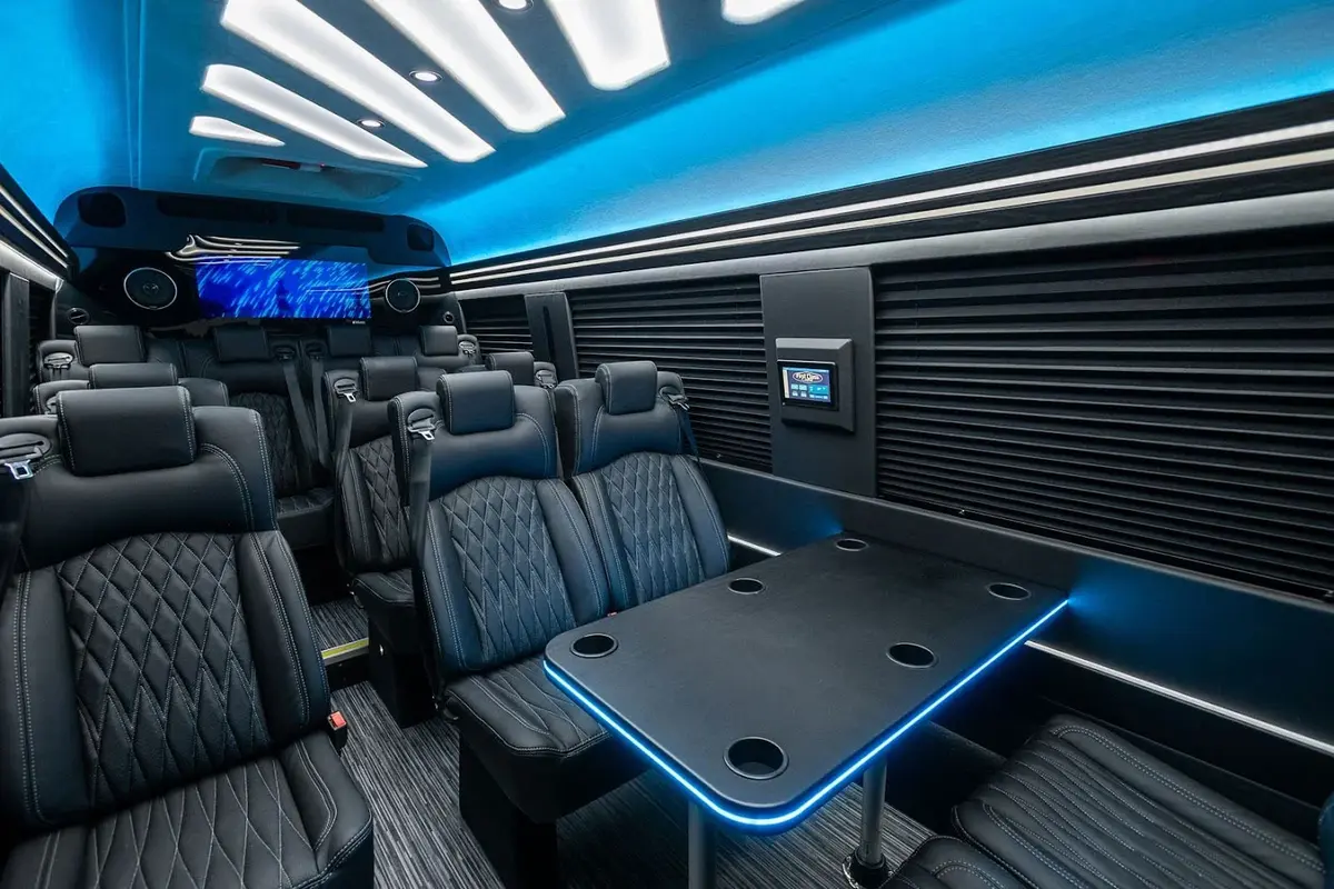 Interior of a luxurious van with black leather seats, a central table, a screen, speakers and a control panel on the side. Curtains closed for privacy
